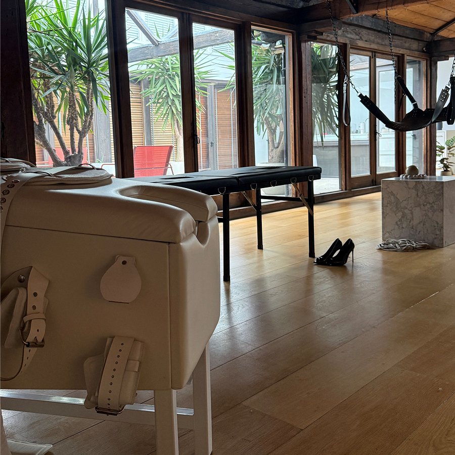 Room with massage tables, wooden floors, large windows, hanging leather swing, and black high heels on floor.