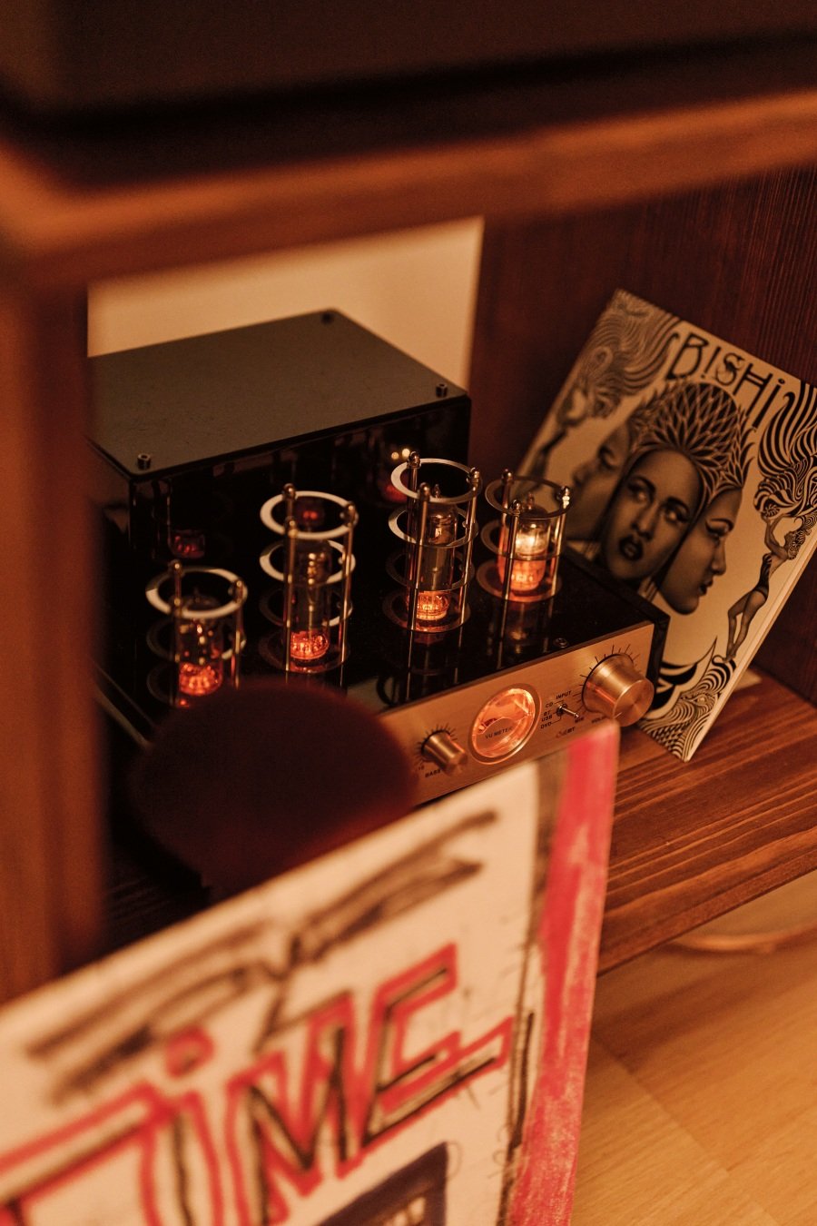 Vintage tube amplifier glowing warmly on wooden shelf with artistic vinyl album covers nearby.