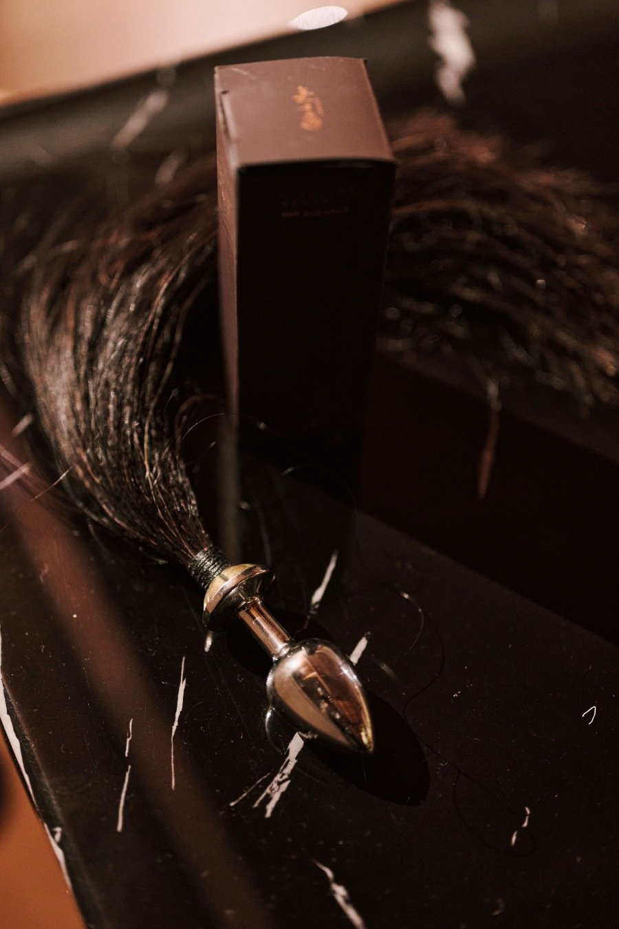 Silver metal butt plug with black tassel placed on black marble surface beside a small dark box.
