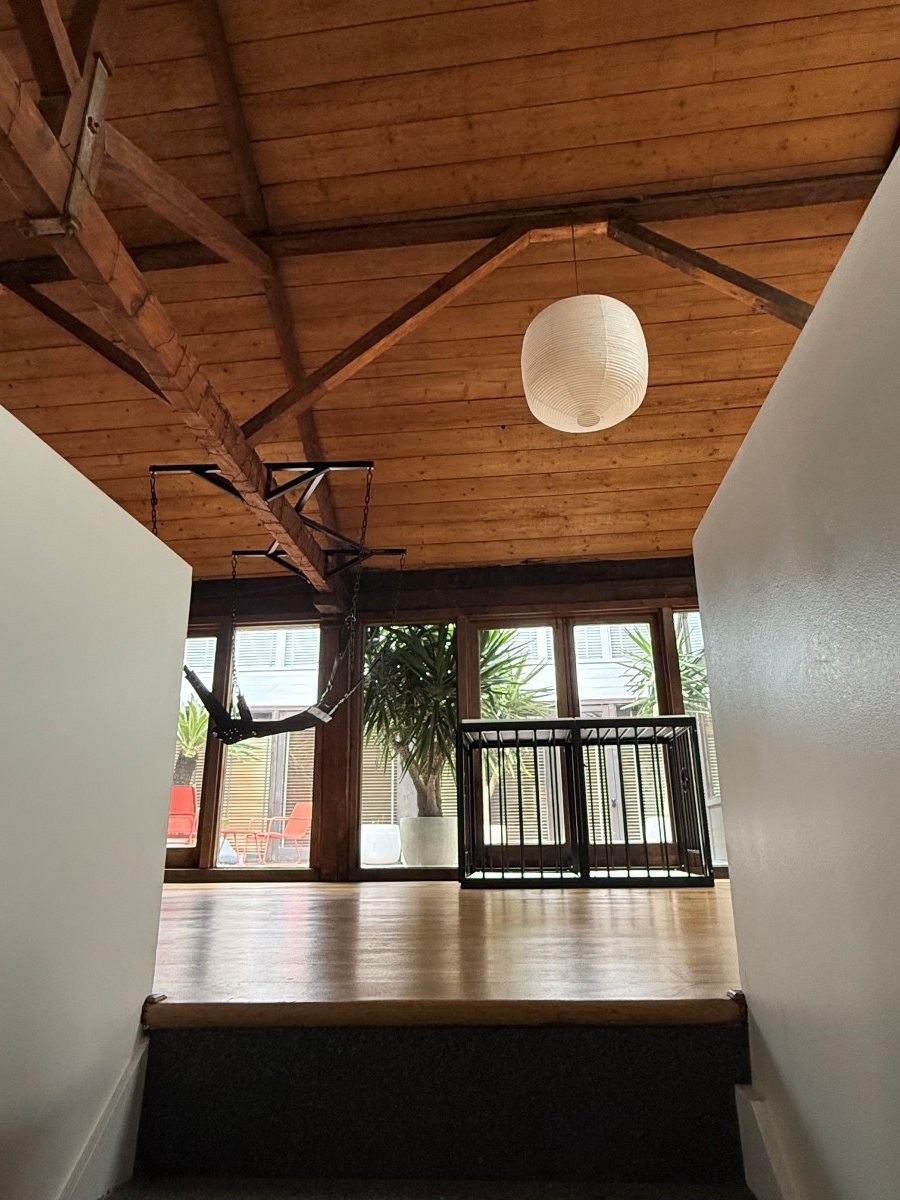 Interior loft space with wooden ceiling, hanging white lantern, large windows, plants, and a black metal cage.
