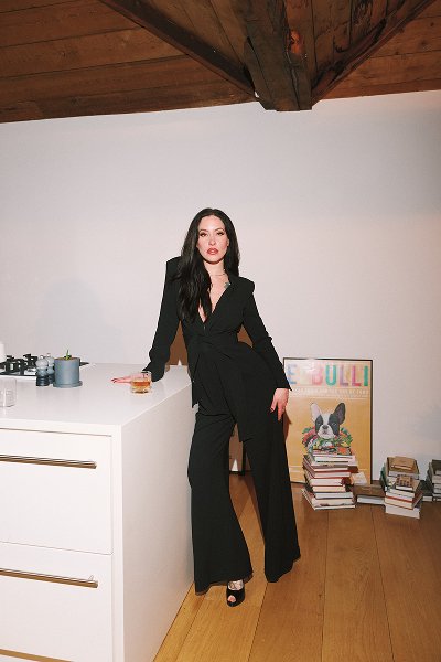 Woman in a black suit leaning on a white kitchen island with stacked books and dog poster behind her.